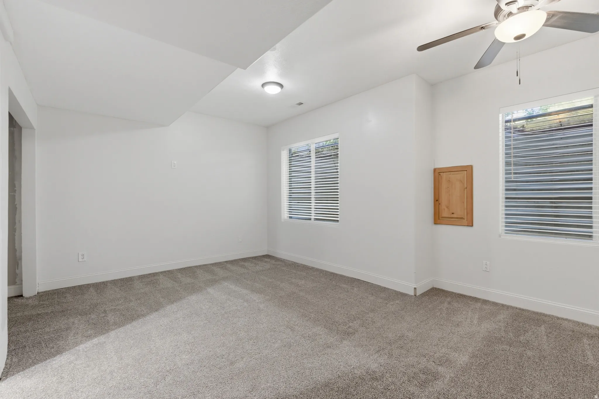 Spacious room featuring light gray carpeting, two windows with horizontal blinds, and a built-in wooden panel