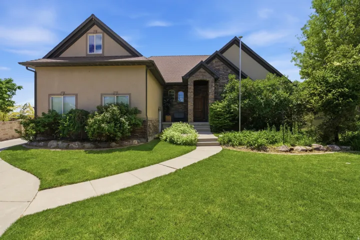 Contemporary stucco and stone-clad residence featuring a gabled roofline, a concrete walkway leading to the entrance, a stone-faced entryway, and established landscaping with mature greenery