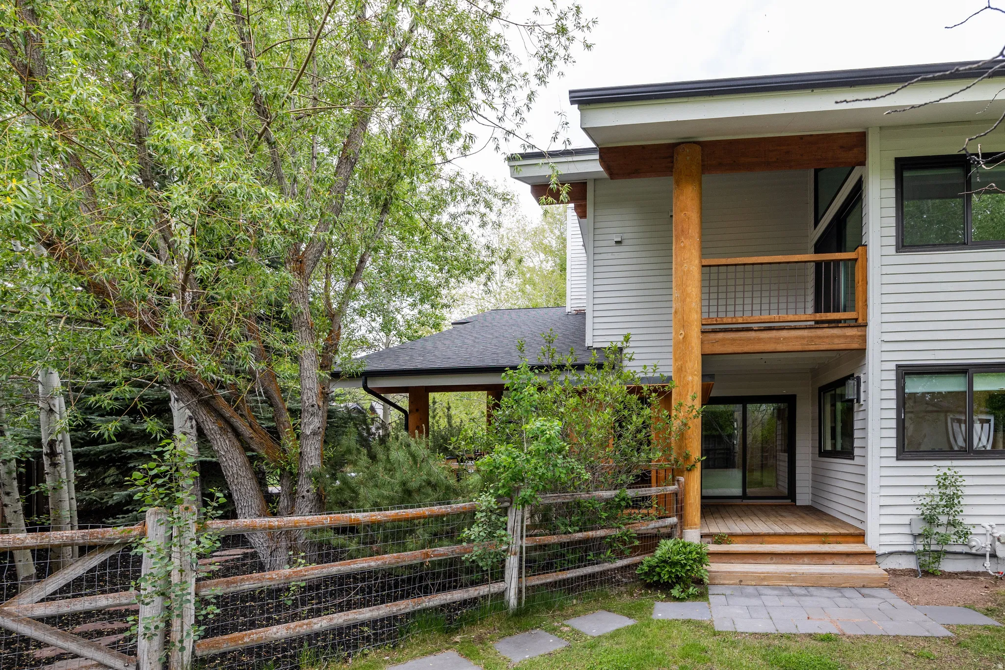 Contemporary exterior featuring light-colored siding, a second-story balcony with wood railing, a ground-level wood deck, black-framed windows, and stone pavers leading to the entrance