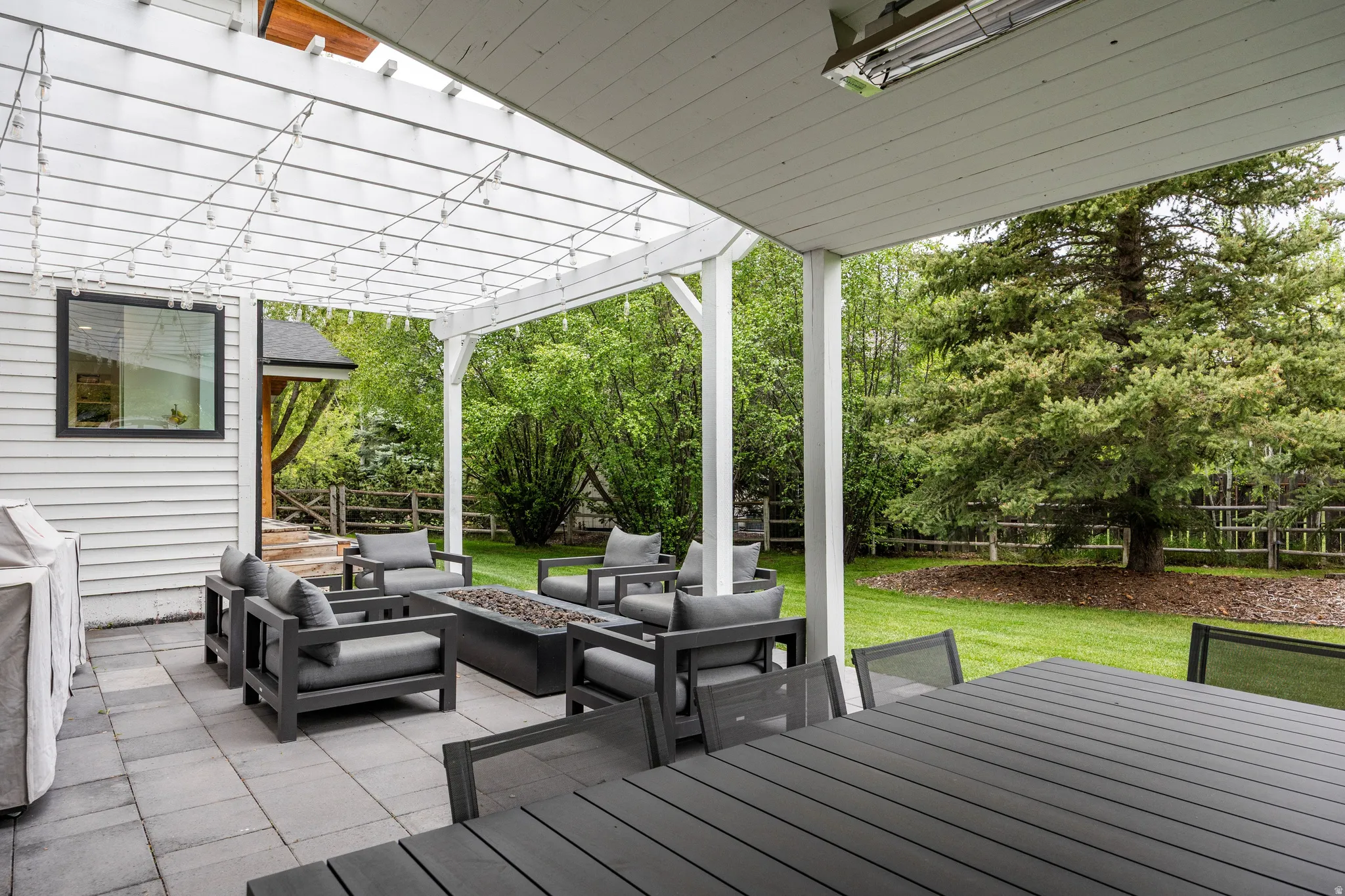 Expansive outdoor patio featuring paver flooring, a white pergola with string lights, and a built-in fire pit
