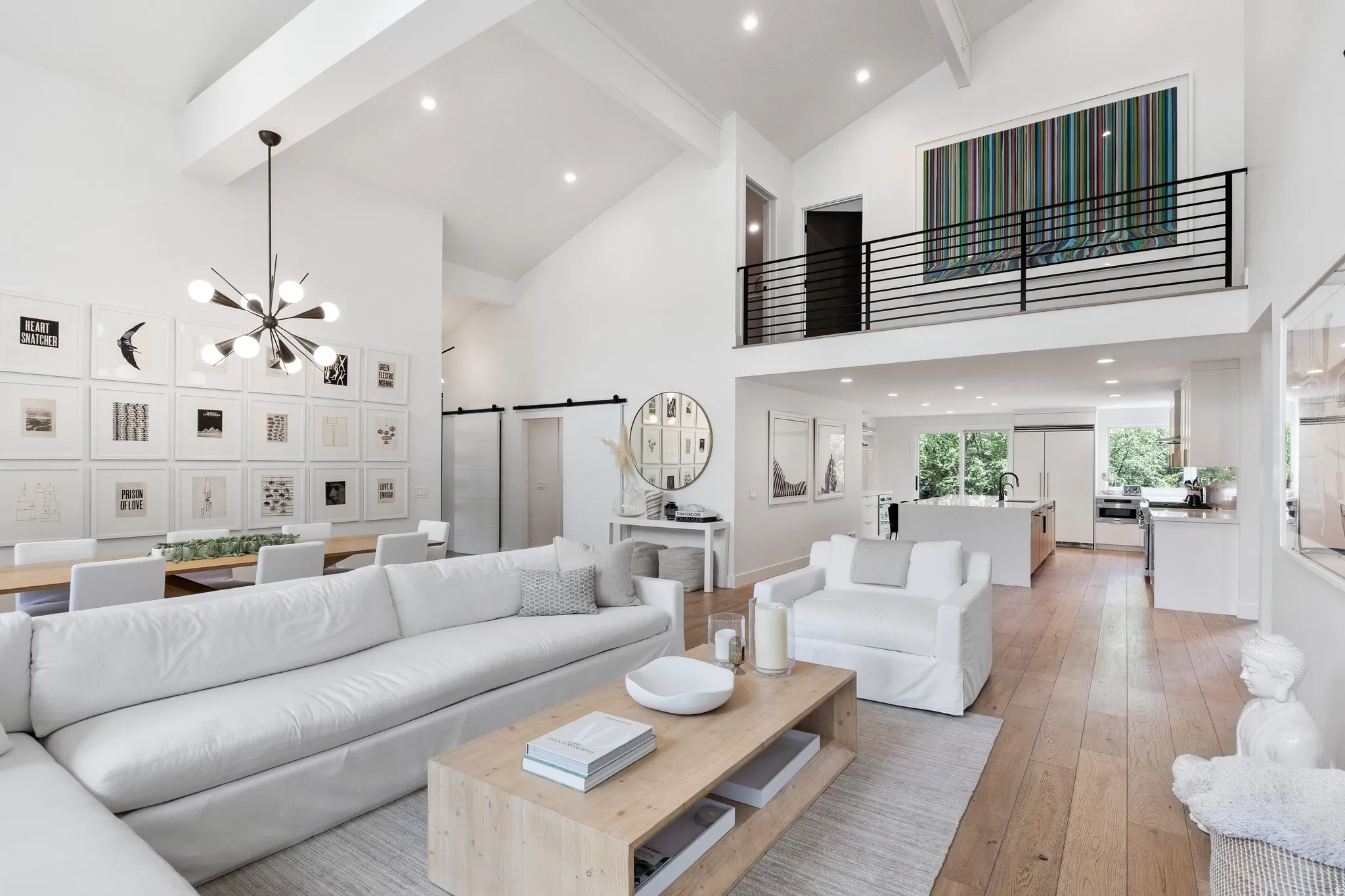 Spacious interior featuring high vaulted ceilings, light wood-finish flooring, a modern chandelier, a kitchen with white cabinetry and stainless steel appliances, and an upper-level balcony with a black metal railing