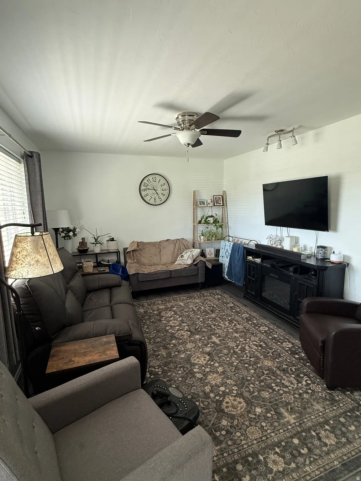 Living area featuring wood-finish flooring, a ceiling fan with integrated lighting, and a window with blinds and drapes