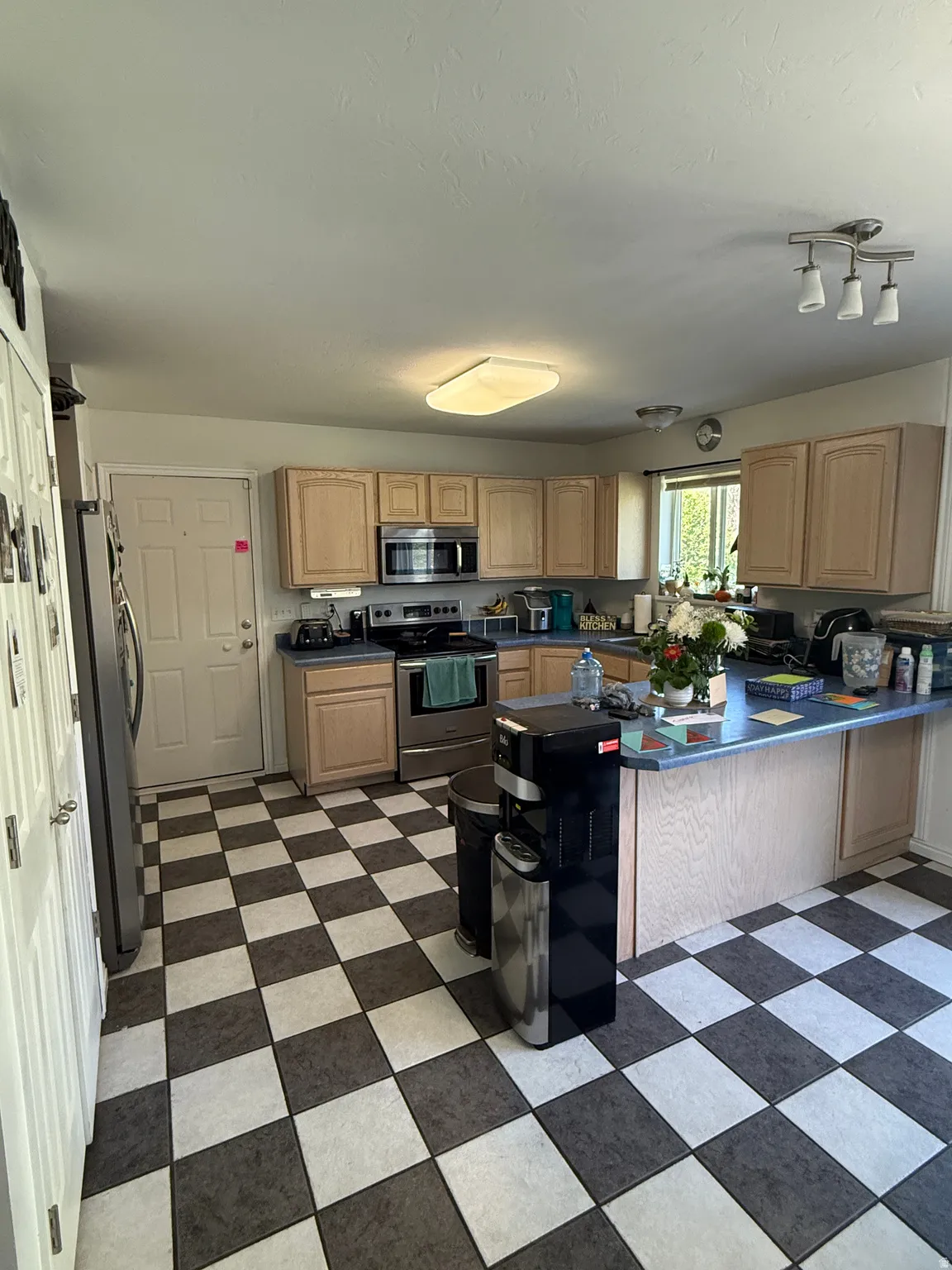 Kitchen featuring checkered flooring, light wood-finish cabinetry, stainless steel appliances, light blue countertops, and a built-in breakfast bar