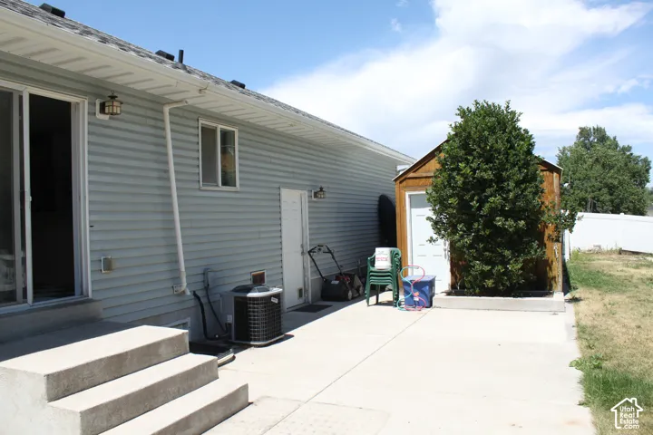 Rear exterior featuring light blue siding, a sliding glass door, a secondary exterior door, and a built-in storage shed with an evergreen shrub