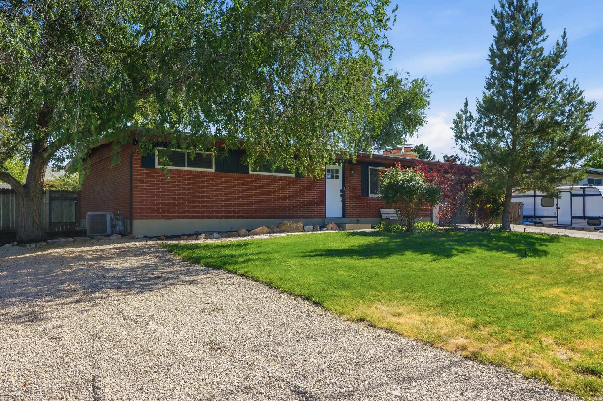 Brick exterior featuring a gravel driveway, mature trees, a manicured lawn, and a white entry door with side shutters