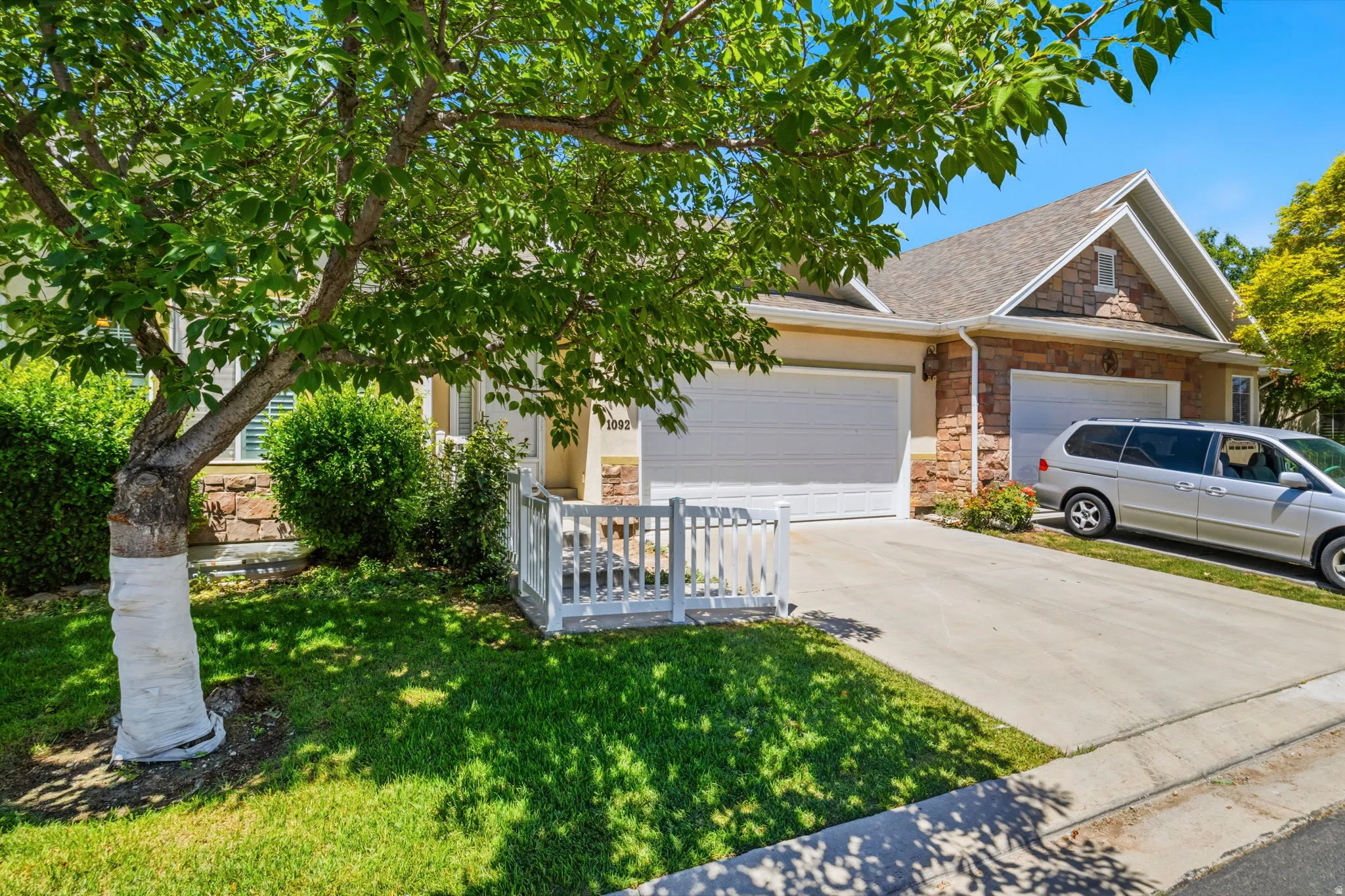 Front exterior featuring a two-car garage, stone and siding facade, concrete driveway, and front lawn with mature tree