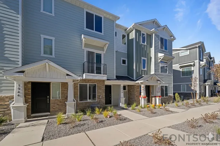 Contemporary townhome exterior featuring light blue horizontal siding, stone veneer accents, a covered entry with white columns, a second-story Juliet balcony, and a multi-pane window