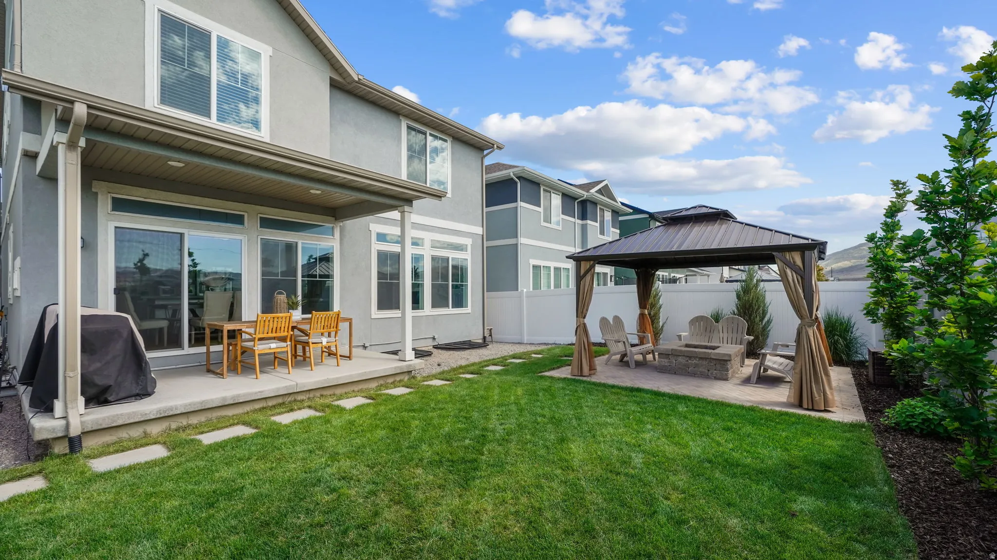 Expansive backyard featuring a covered patio, manicured lawn, and a paved seating area with a built-in fire pit