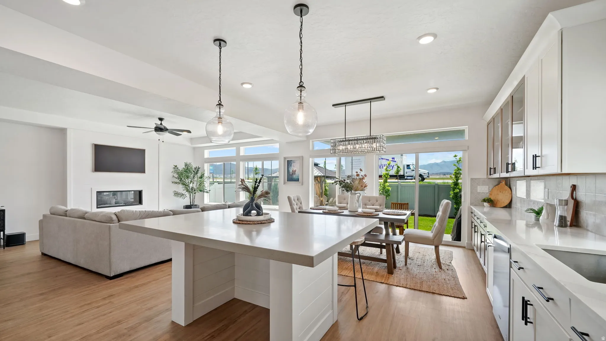 Open-concept living space featuring wood-finish flooring, a kitchen island with pendant lighting, white cabinetry with black hardware, a tiled kitchen backsplash, and large sliding glass doors