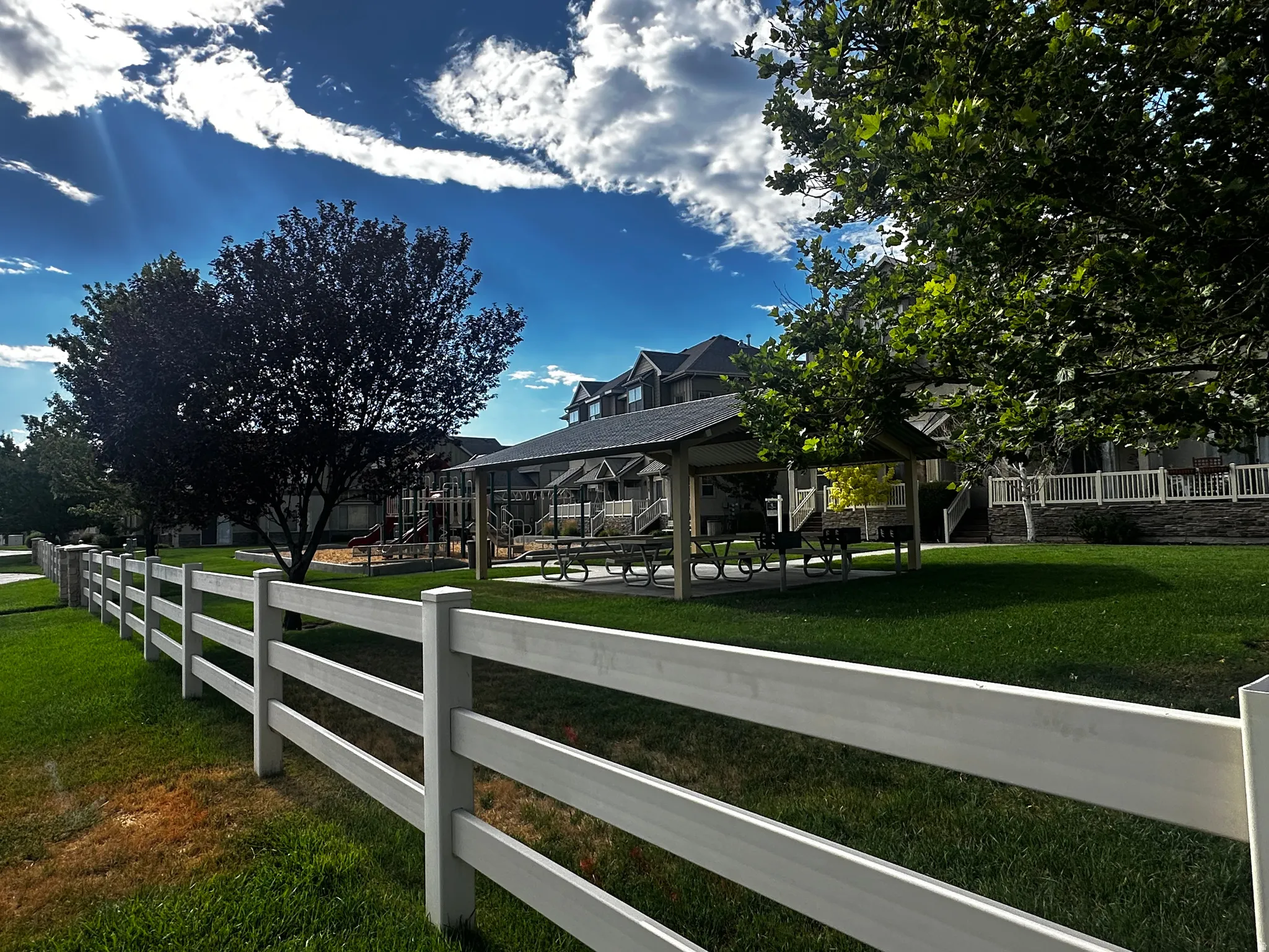 Community green space featuring a white split-rail fence, a shaded picnic area with tables, a playground, and mature trees