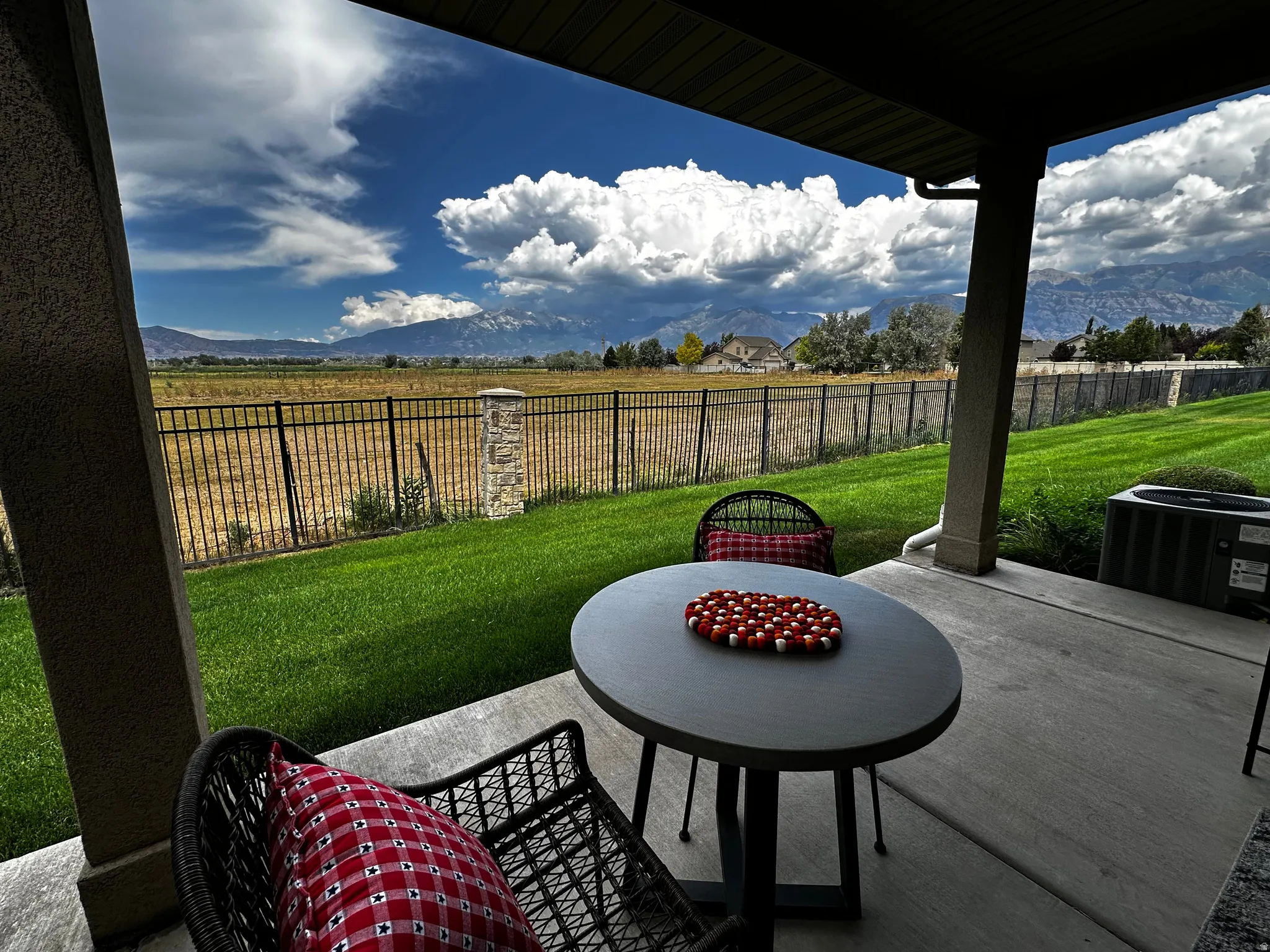 Covered patio with concrete flooring, framed by a manicured lawn and black metal fencing, offering a mountain backdrop