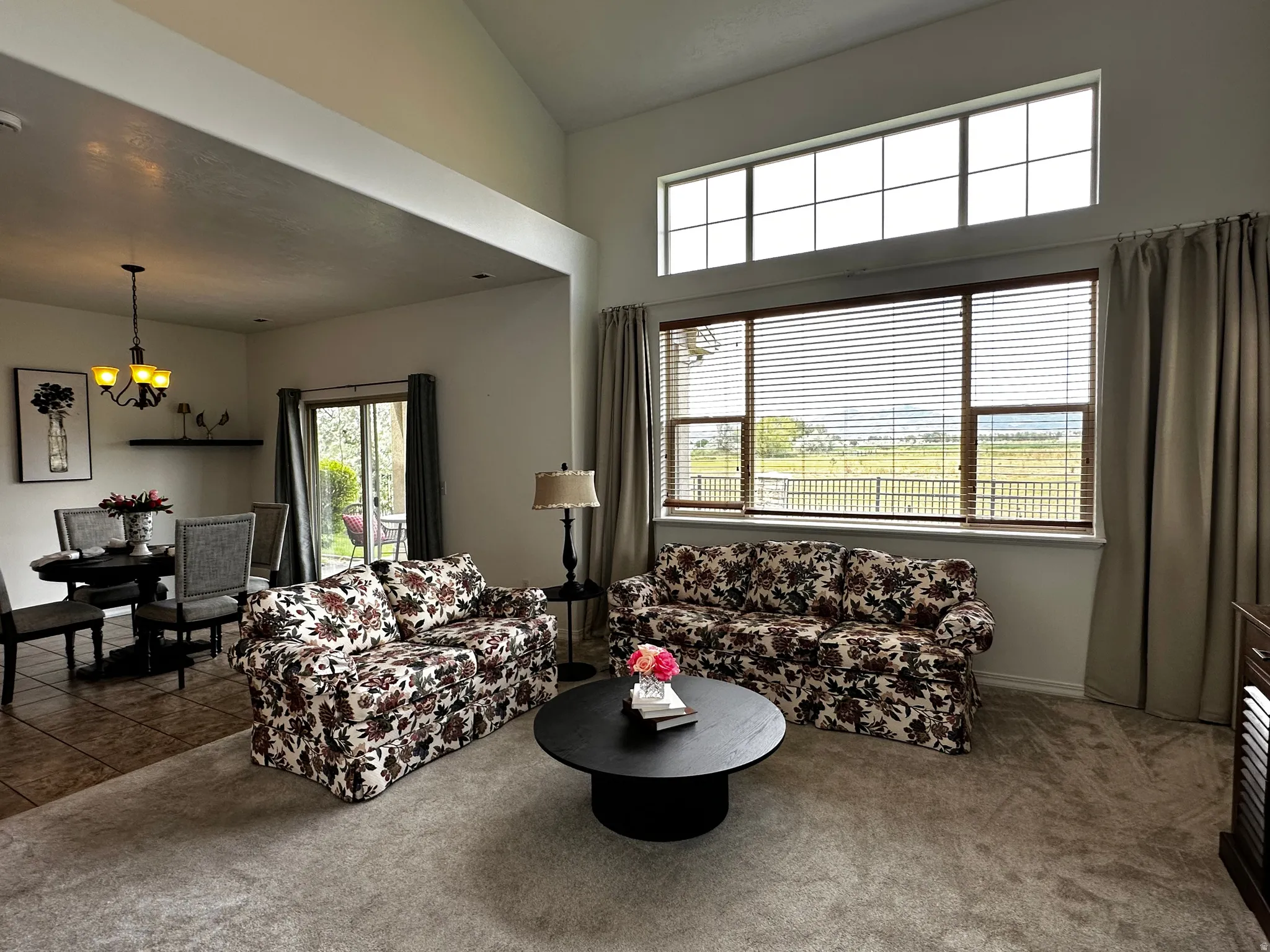 Open-concept living area featuring vaulted ceilings, a large picture window with horizontal blinds, and an upper transom window