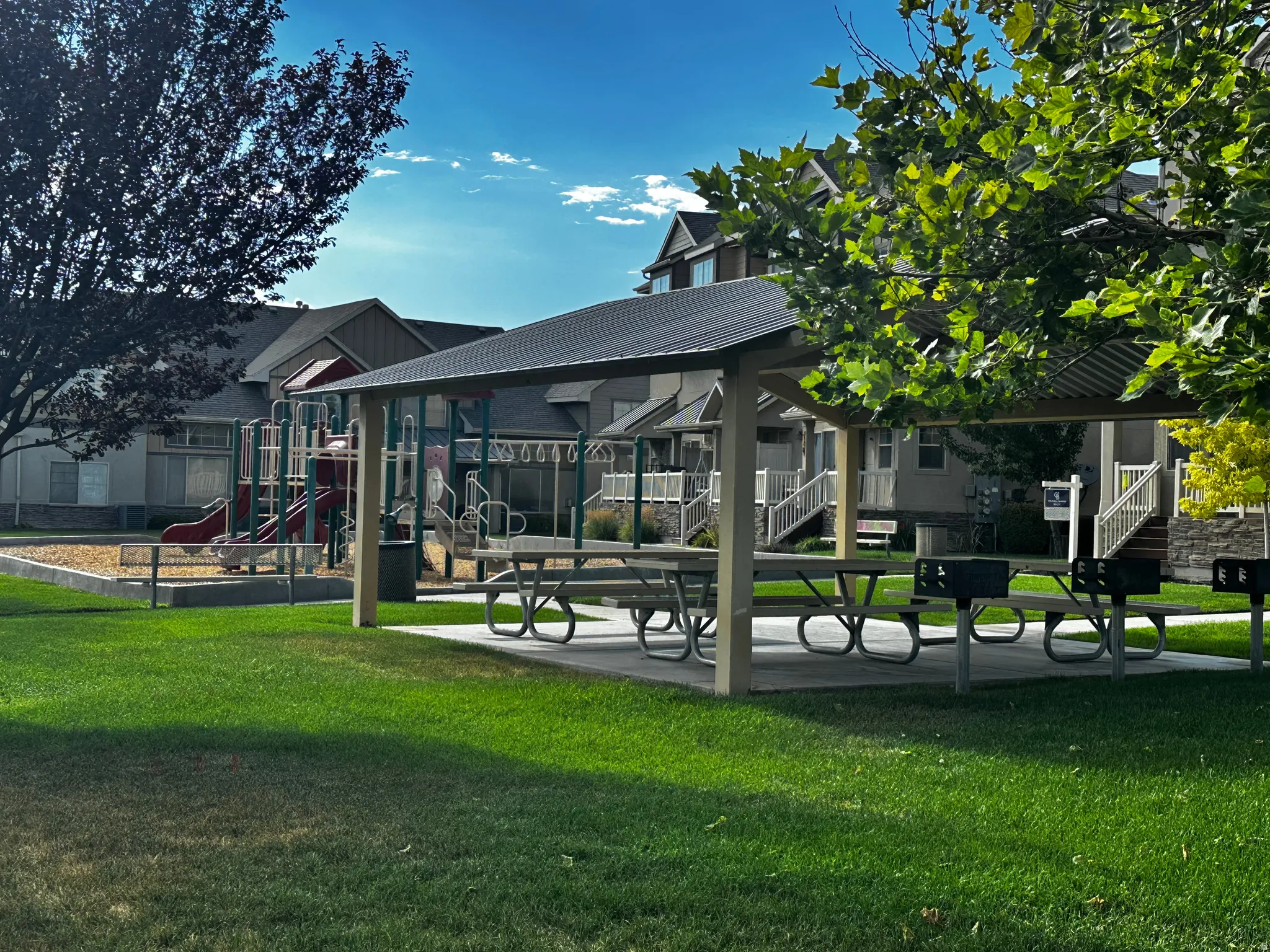 Community park area featuring a covered picnic pavilion with multiple tables, paved grilling stations, a playground with slides and swings, and manicured green space