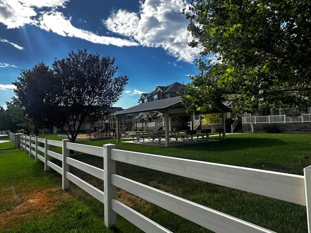 Community green space featuring a white split-rail fence, a shaded picnic area with tables, a playground, and mature trees