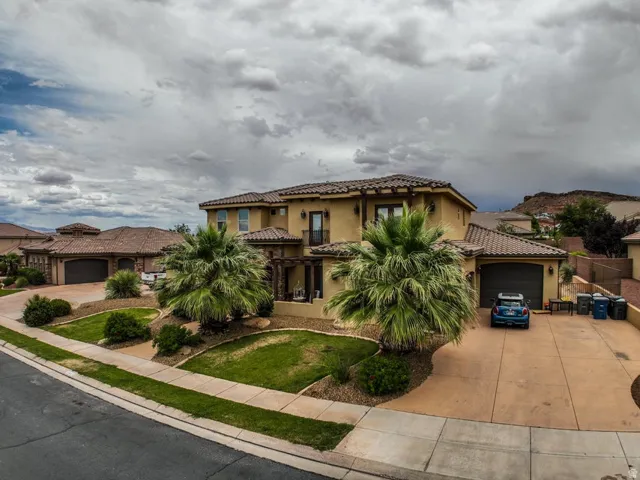 Stucco exterior with a tile roof, featuring a two-car garage, mature palm trees, and a decorative pergola