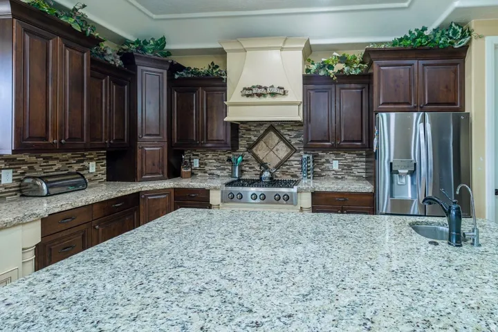 Spacious kitchen featuring a large island with an undermount sink, dark wood cabinetry, light-toned granite countertops, a tiled backsplash, and stainless steel appliances