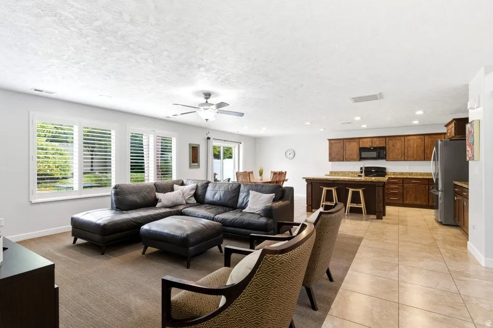 Open-concept living area featuring a kitchen with wood cabinetry and a central island, tile flooring, carpeted living room, and multiple windows with plantation shutters
