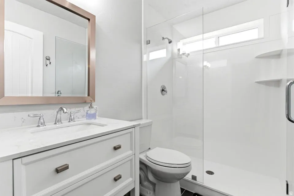 Bathroom featuring a single vanity with a white countertop and rectangular undermount sink, a framed mirror, and a glass-enclosed shower with white paneling and a transom window