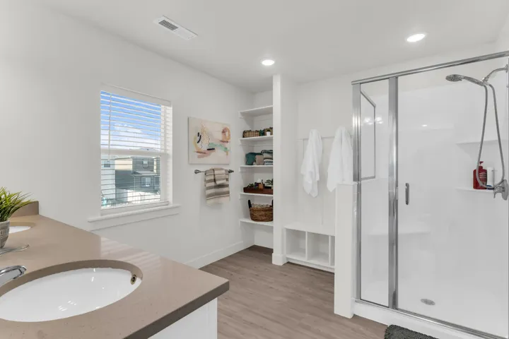 Spacious primary bathroom featuring a dual vanity with undermount sinks, a window with blinds, built-in shelving, and a glass-enclosed shower