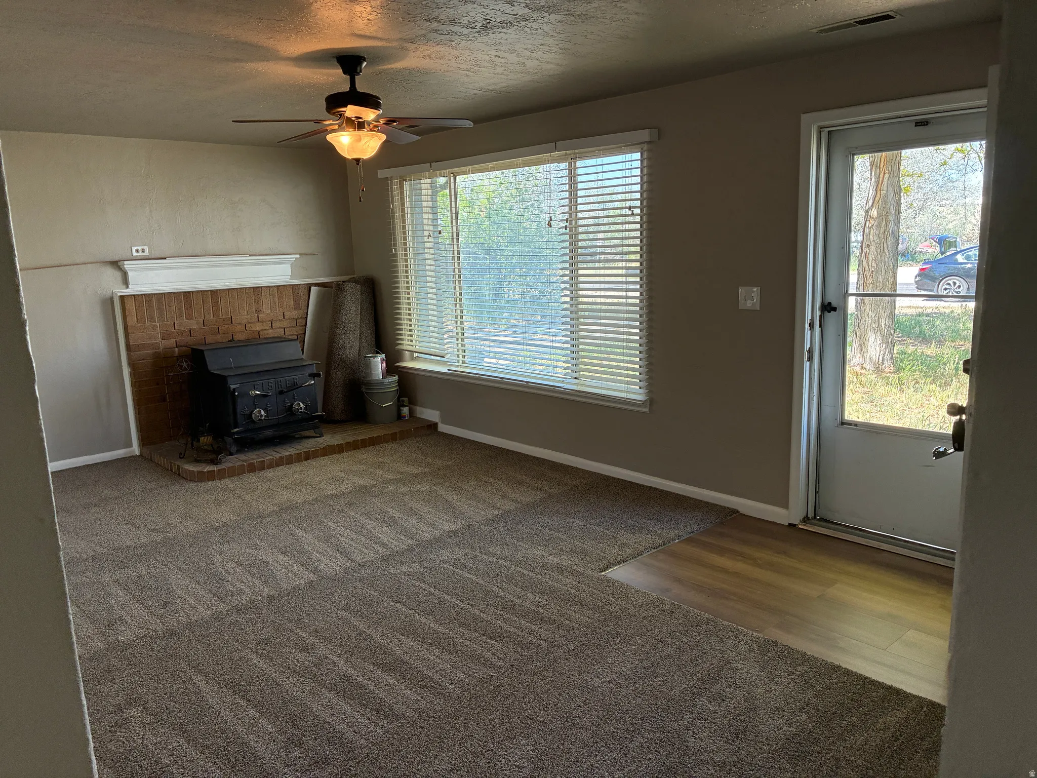 Living area featuring an exposed brick fireplace with a black wood-burning stove, a ceiling fan with integrated lighting, a large window with horizontal blinds, and a combination of carpet and wood-finish flooring