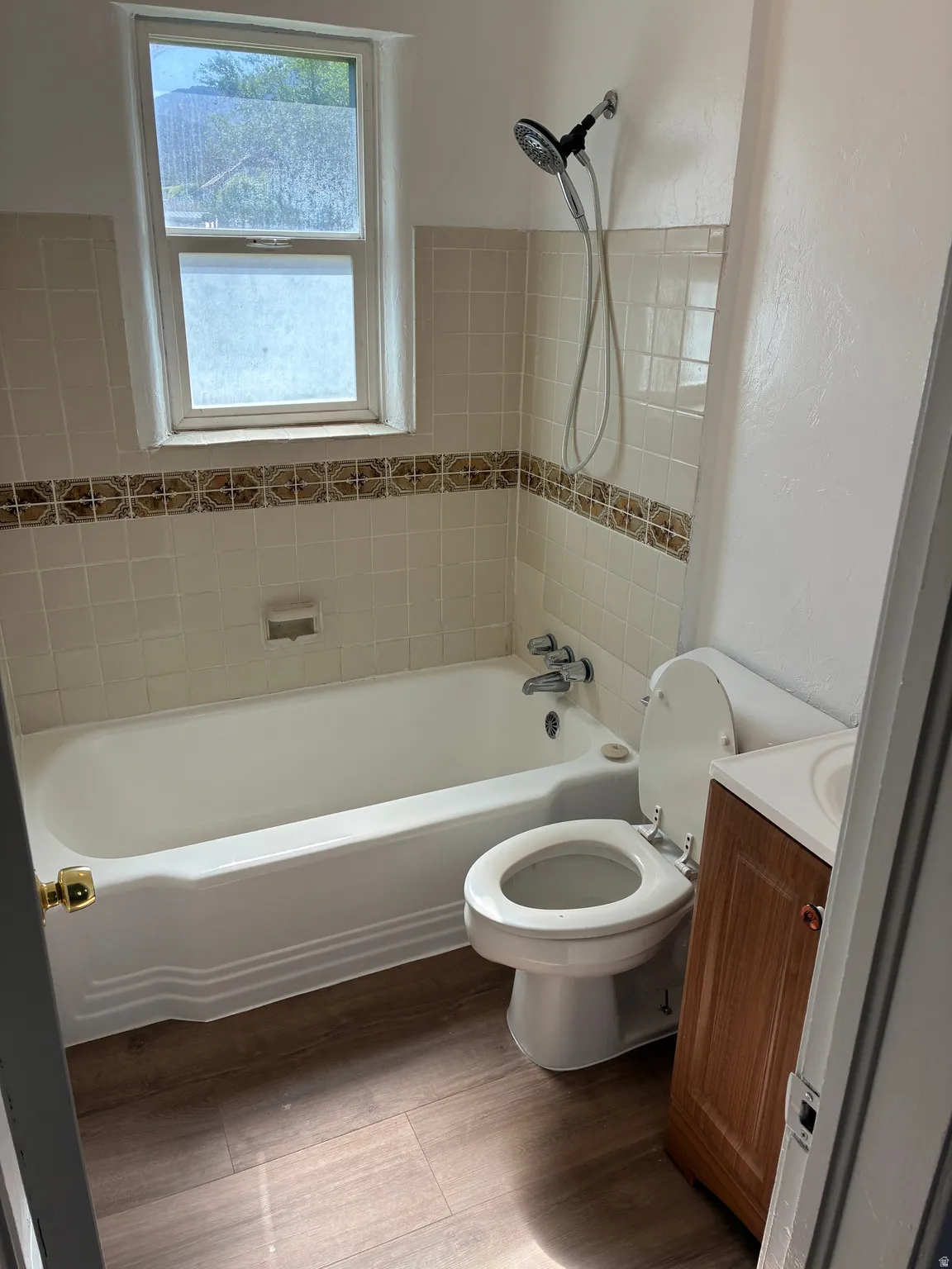 Bathroom featuring a white bathtub, ceramic wall tiles with a decorative accent band, wood-finish flooring, a window, and a vanity with a white countertop