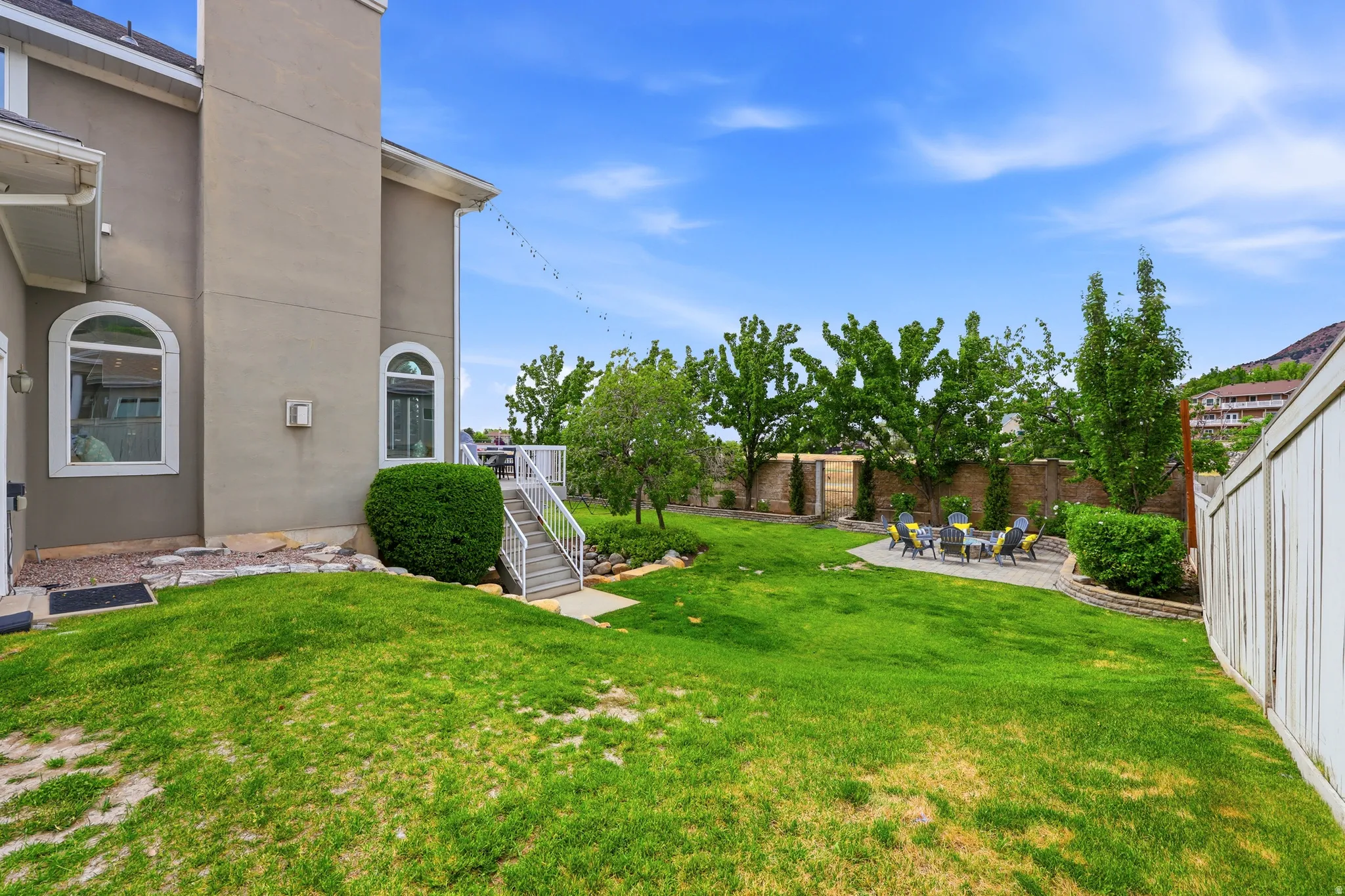 Expansive backyard featuring a concrete patio, mature trees, manicured lawn, and a white privacy fence