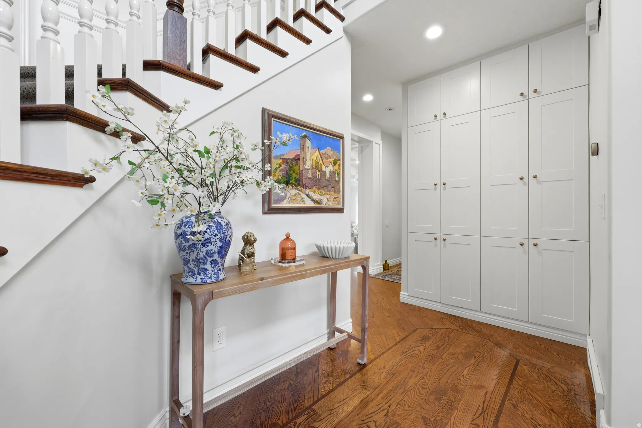 Hardwood flooring with a geometric inlay pattern, built-in white paneled cabinetry, staircase with white balusters and dark wood treads, white wall finishes, recessed lighting
