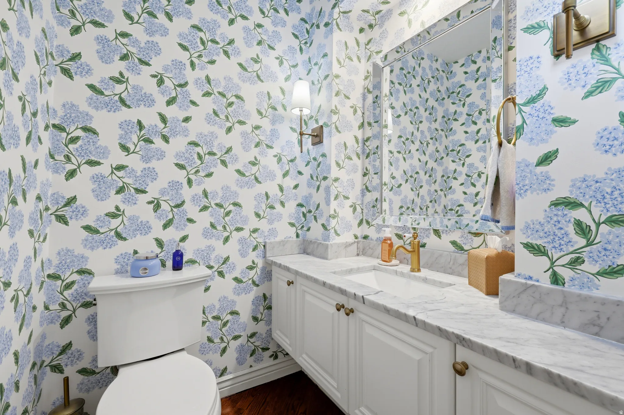 Powder room featuring floral wallpaper, a white vanity with paneled doors, brass hardware, a white undermount sink, and a marble countertop