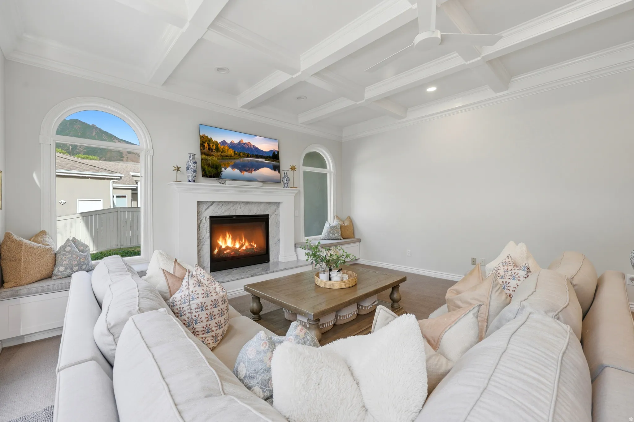 Living area featuring a gas fireplace with a marble surround and white mantel, coffered ceilings with recessed lighting, wood-finish flooring, and arched windows with integrated window seating