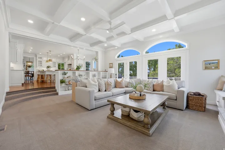 Spacious living area featuring coffered ceilings, carpet flooring, built-in shelving, and direct access to an elevated kitchen area with wood-finish flooring