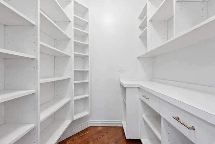 Custom storage area featuring extensive white built-in shelving, white countertops, multiple drawers with brass-finish pulls, and wood-finish flooring