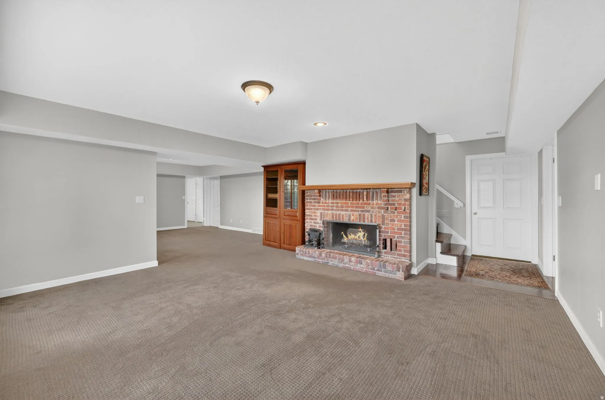 Spacious recreation room featuring a brick fireplace with a wood mantle, an integrated wood-finish display cabinet, recessed lighting, and access to a stairwell