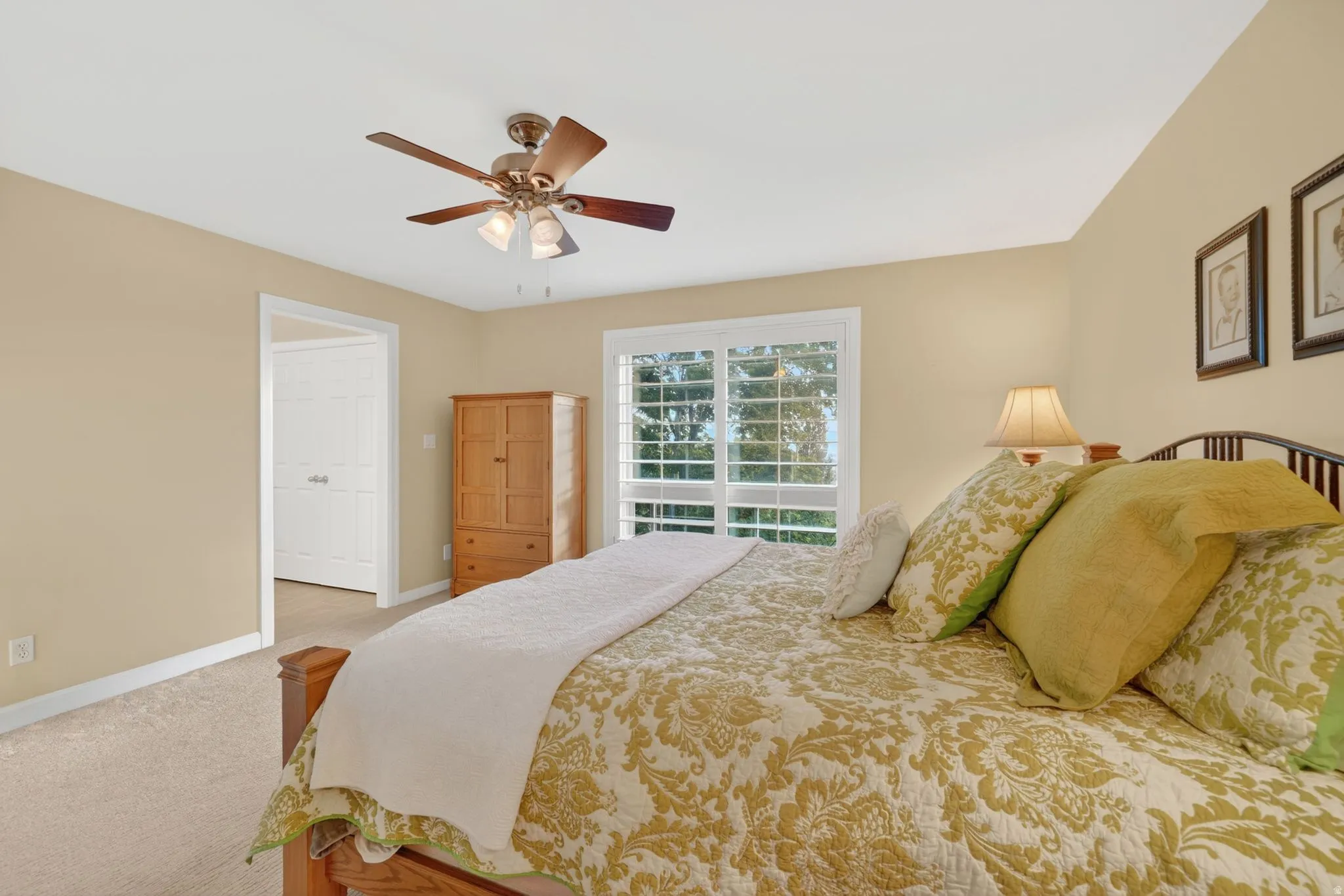 Carpeted bedroom featuring a ceiling fan with light kit, a large window with white plantation shutters, and an interior door with white trim
