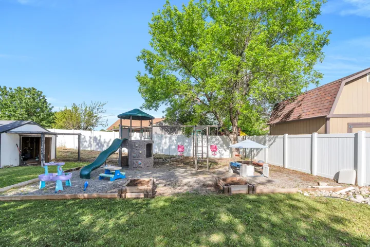 Expansive backyard featuring a large tree, a white privacy fence, a storage shed, a play structure with a slide, and a gravel area