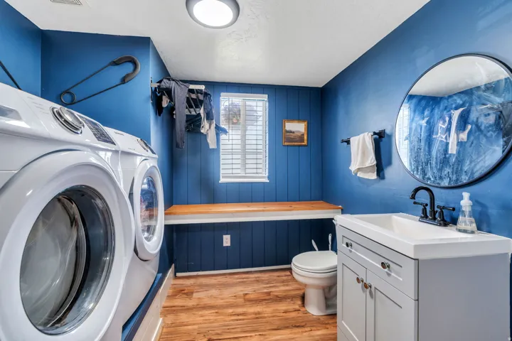 Utility room featuring wood-finish flooring, a white vanity with an integrated sink, a round wall-mounted mirror, and a window with horizontal blinds