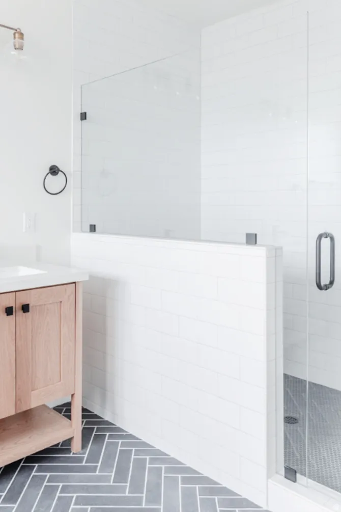 Contemporary bathroom featuring a light wood-finish vanity with black hardware, white subway tile walls, a frameless glass shower enclosure, and grey herringbone-patterned floor tile