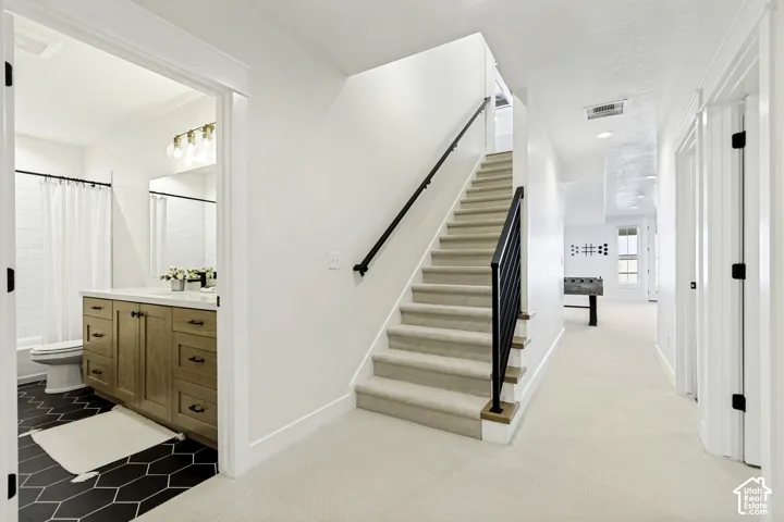 Carpeted staircase with black iron railing, featuring a lower-level hallway with recessed lighting and a visible bathroom entrance