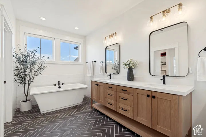 The primary bath features a freestanding soaking tub with a wall-mounted faucet, herringbone tile flooring, dual sink vanity with wood cabinetry, white countertops, and stylish black-framed mirrors with brass light fixtures