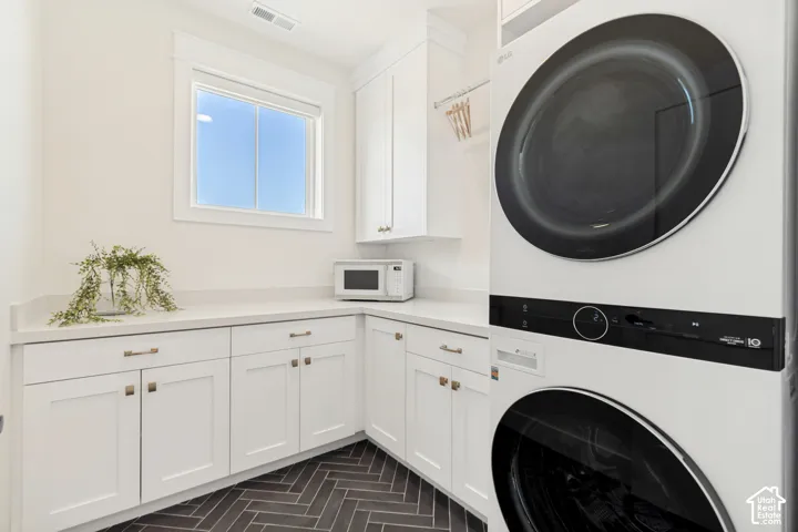Modern laundry area featuring a stacked washer and dryer, extensive white cabinetry with bronze hardware, light-toned countertops, and dark herringbone tile flooring