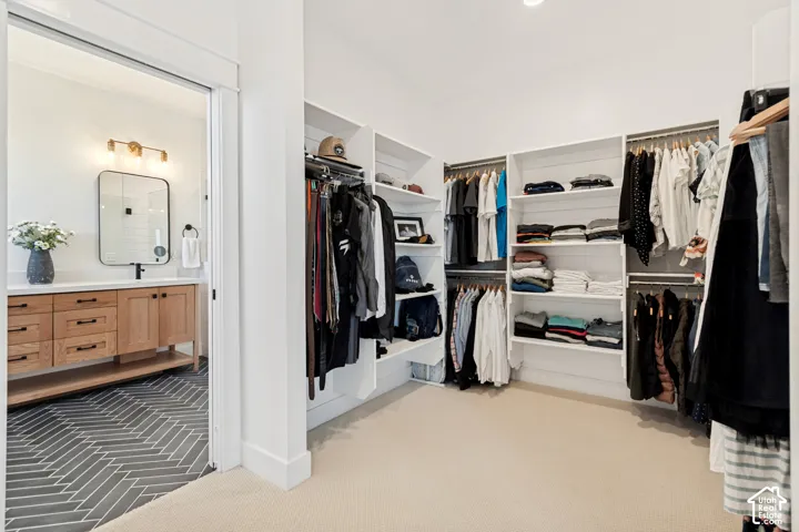 Walk-in closet featuring extensive built-in shelving, multiple hanging rods, and neutral carpeting, with a clear sightline into an adjacent bathroom featuring a wood-finish vanity, white countertop, and patterned tile flooring
