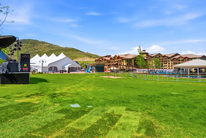 Expansive green lawn surrounded by a white picket fence, with a large event tent and multi-story buildings in the background
