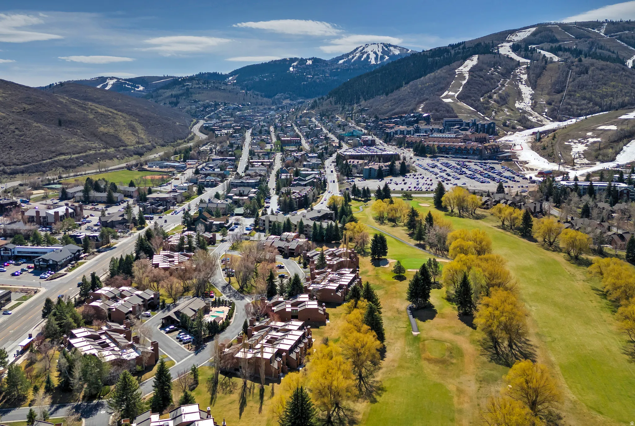 Expansive aerial showcasing a mountain town with snow-capped peaks, surrounding green hillsides, a golf course with mature trees, and numerous residential and commercial buildings