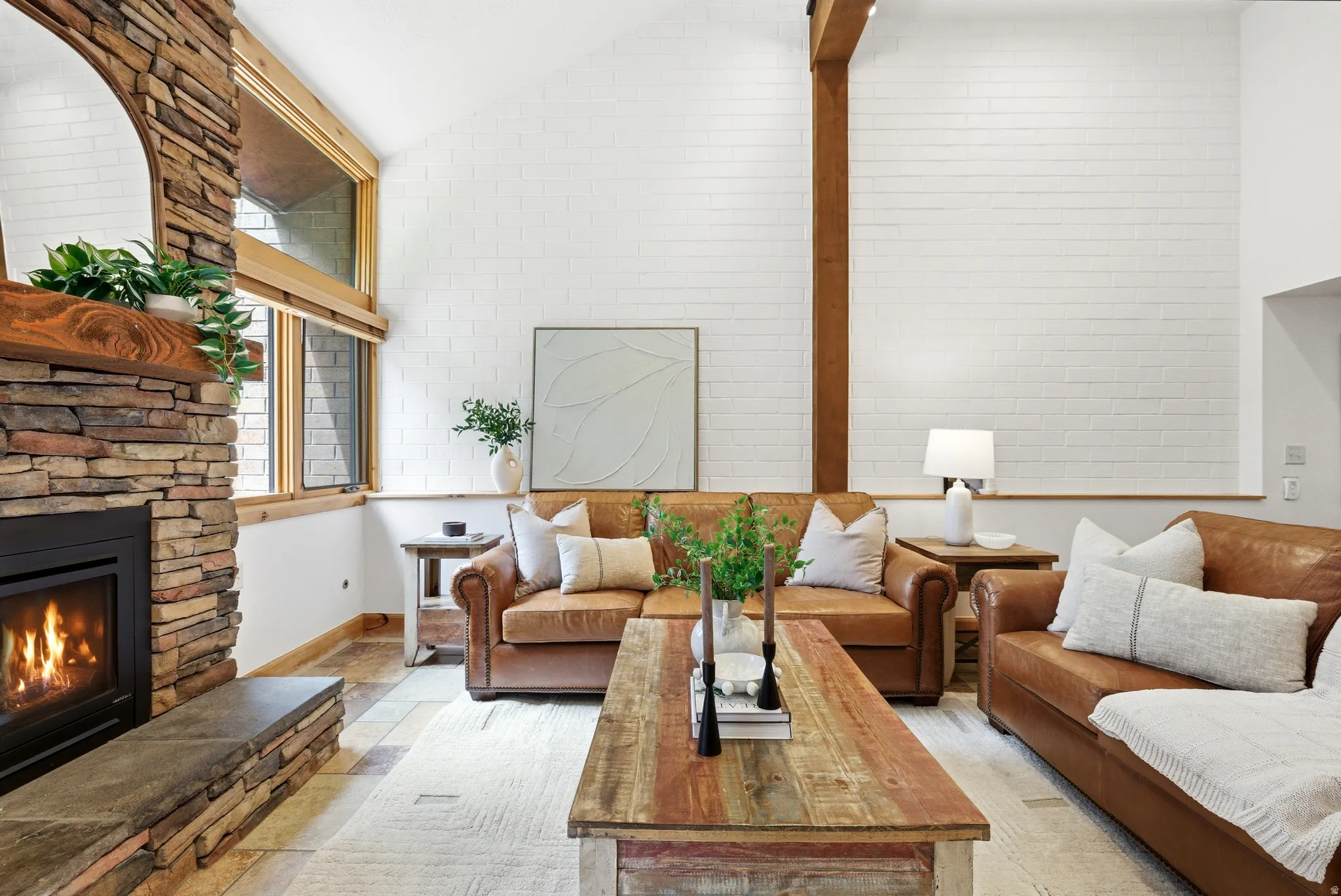 Living area featuring a floor-to-ceiling stone fireplace with a wood mantel, vaulted ceiling, exposed wood beam, white brick accent wall, and wood-frame windows