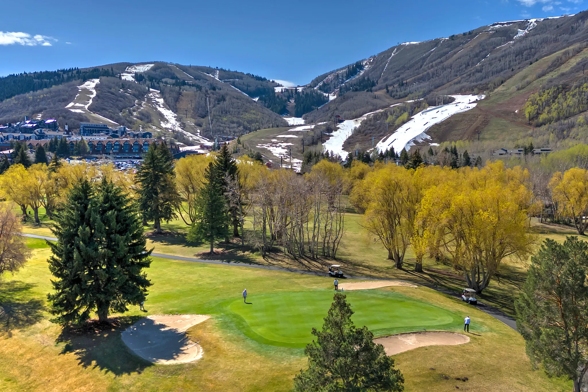 Sprawling golf course green with sand traps and mature trees, set against a backdrop of distant snow-capped mountains and a residential area