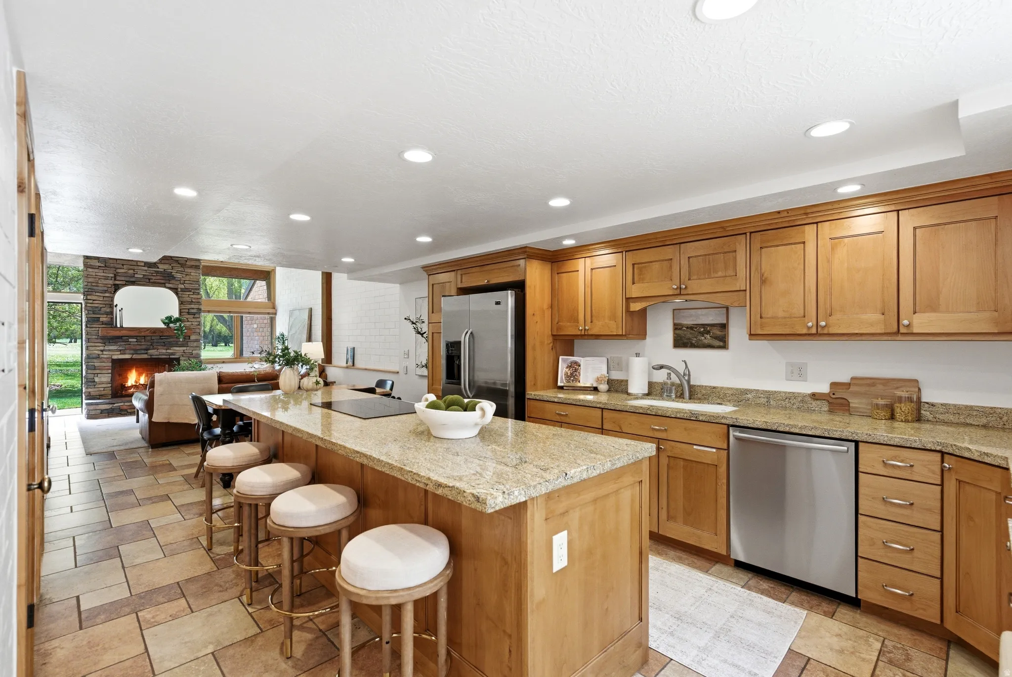 Kitchen and living area featuring a stone fireplace, wood cabinetry, granite countertops, stainless steel appliances, and tile flooring