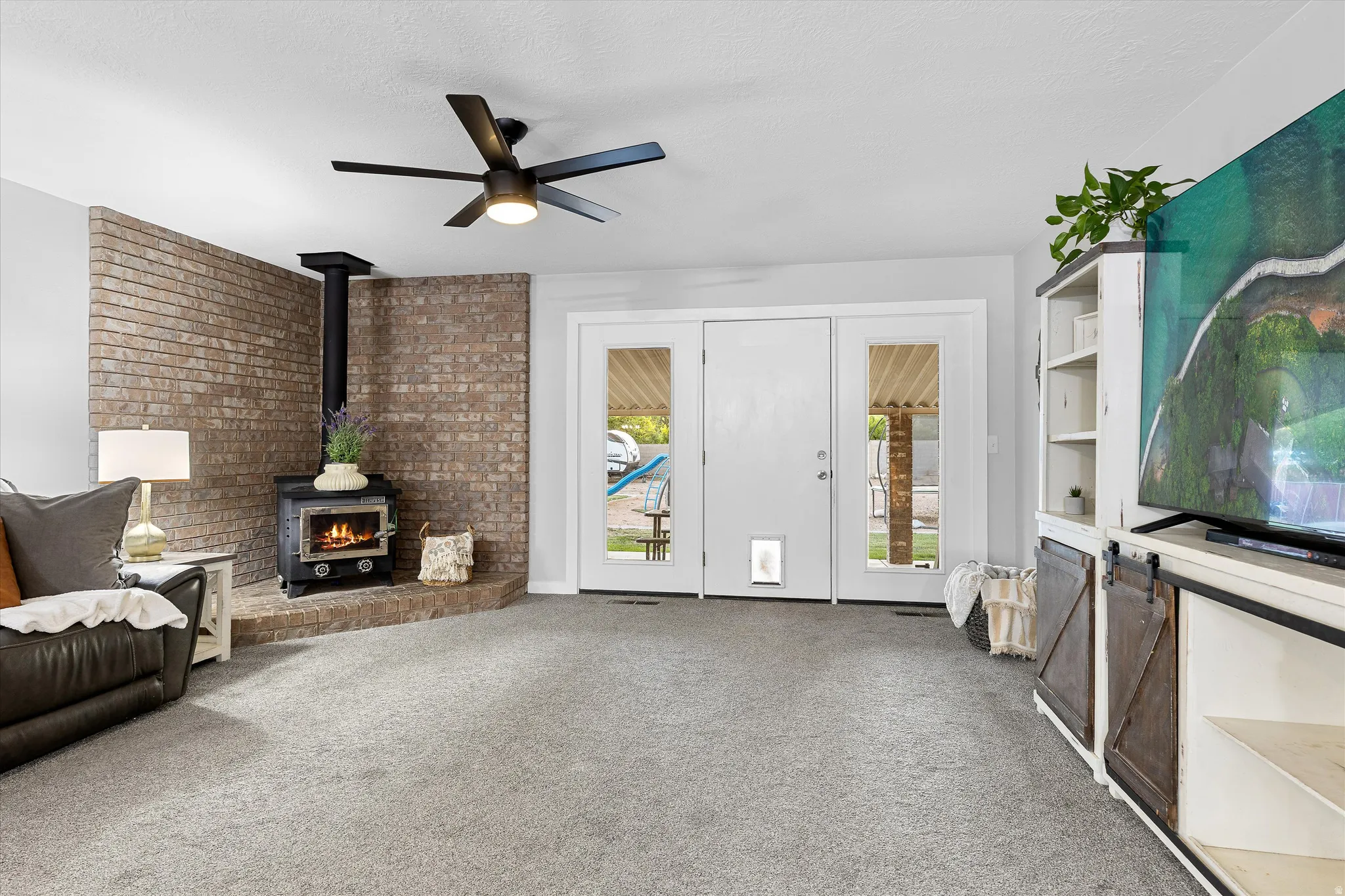 Living Room with Brick-Wall Fireplace. Living area featuring a brick accent wall with a wood-burning stove, gray carpet, and white French doors