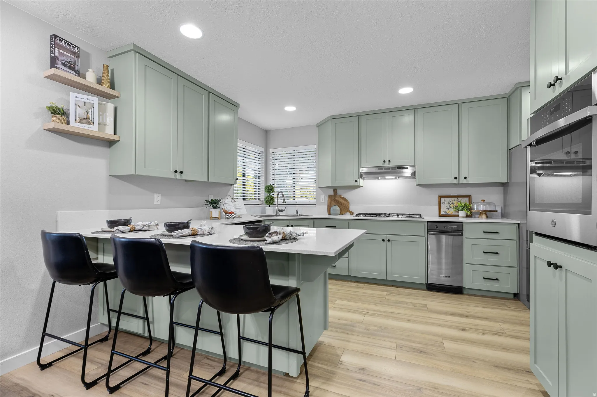 Updated Kitchen with Sage Cabinetry & Island. Kitchen featuring light green cabinetry, a white countertop island with seating, stainless steel appliances, wood-finish flooring, and floating wall shelves