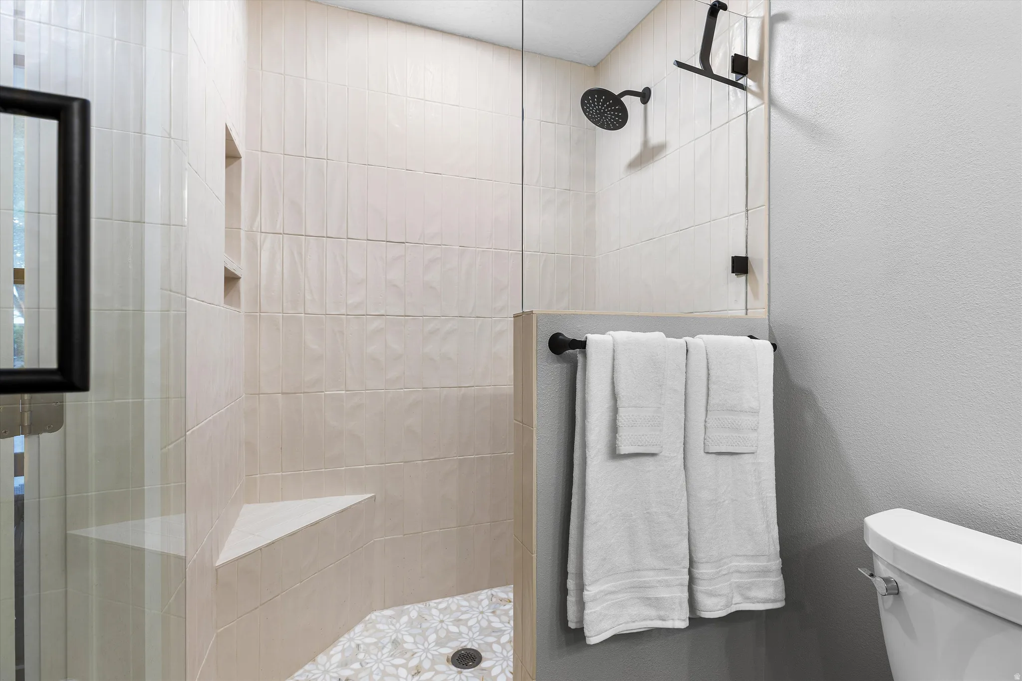 Tiled Walk-In Shower in Primary Bath. Glass-enclosed shower with vertical light-toned tile, integrated wall niche, corner bench seat, and patterned pebble floor