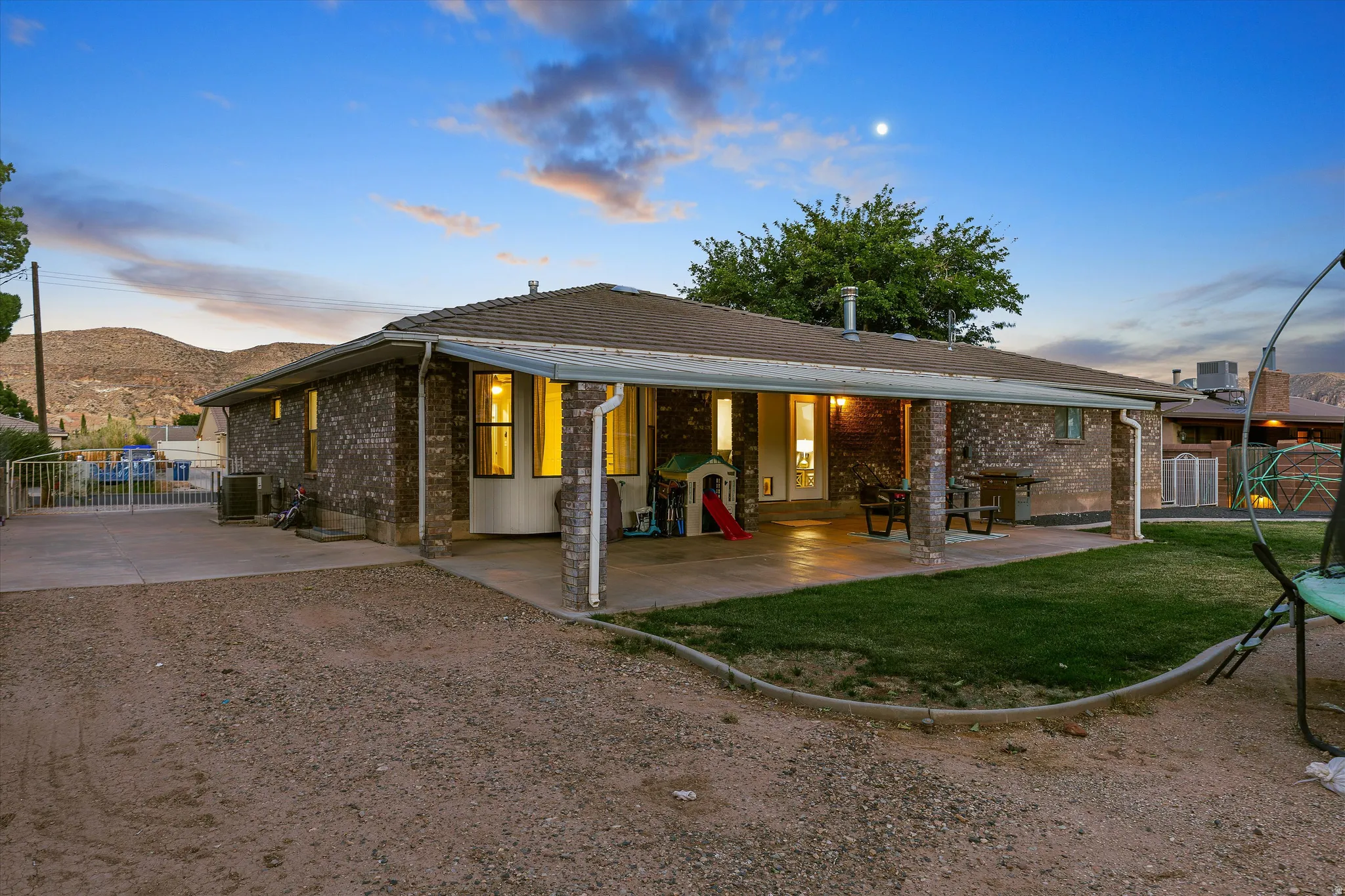 Back of Home at Sunset. Brick exterior ranch-style property featuring a covered patio, a manicured lawn, and a graveled drive