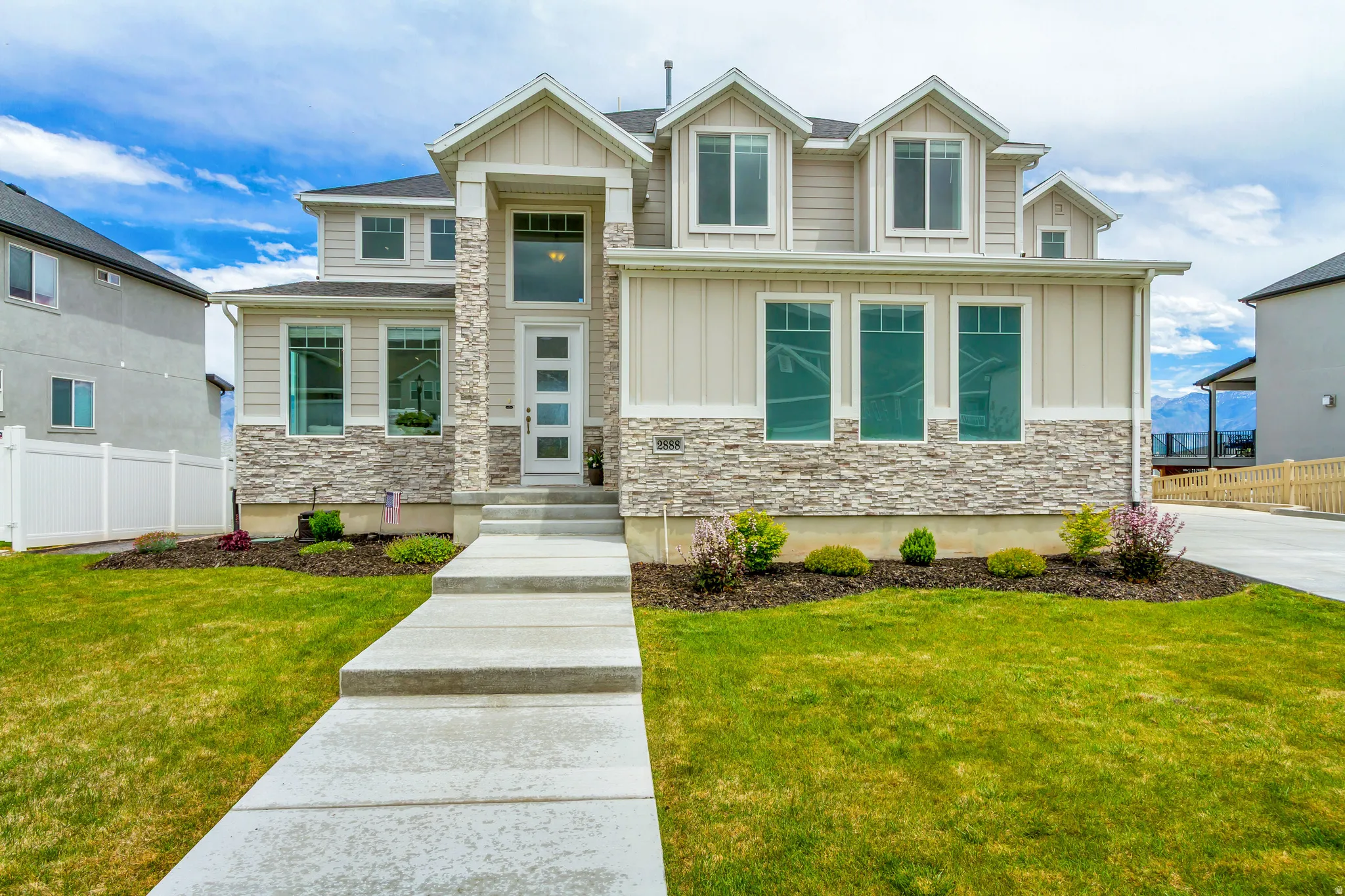Contemporary two-story home featuring a stone and board-and-batten facade, a prominent entry porch with a full-height window, multiple dormer windows, a concrete walkway, a manicured lawn and drip lines in the flower beds.